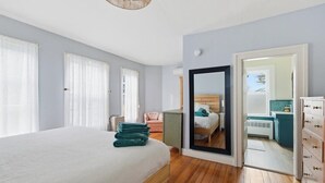 Ein Schlafzimmer mit einem großen Bett, einem Spiegel, einem Sessel und Blick ins Badezimmer durch eine offene Tür.