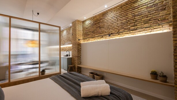 A modern bedroom with a large bed, a wooden-framed sliding door, a brick accent wall, and a small kitchenette.