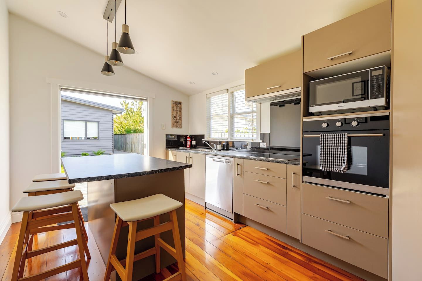 A modern kitchen with a large island, wooden stools, a built-in oven, and a view of the outside.