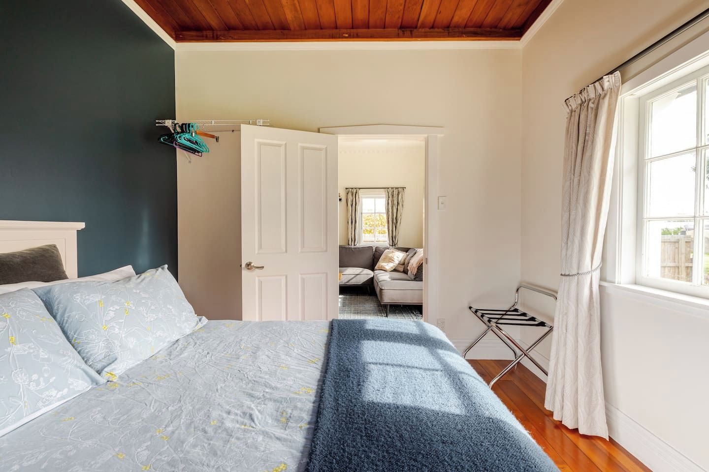 A bedroom with a bed, a door leading to another room, a window with curtains, and a drying rack.
