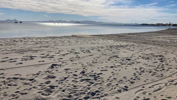 Een strand met voetafdrukken en boten in het water.