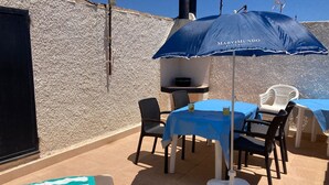 Een terras buiten met een tafel, stoelen, een blauwe parasol en een barbecue.