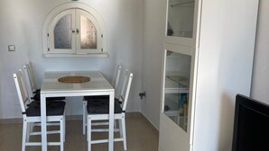 Een kleine, goed verlichte kamer met een witte tafel en stoelen, een klok aan de muur en een kast met glazen deuren.