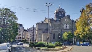 Un bâtiment historique doté d’un dôme imposant et de fenêtres en arc de cercle, entouré de bâtiments modernes et donnant sur une rue bordée de voitures garées.