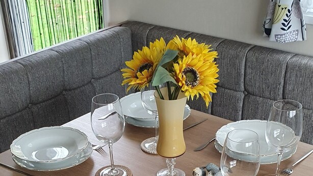 A dining area with a table set for four, a vase with sunflowers, and a grey sofa in the background. - Pet friendly Static caravan. (Dymchurch)