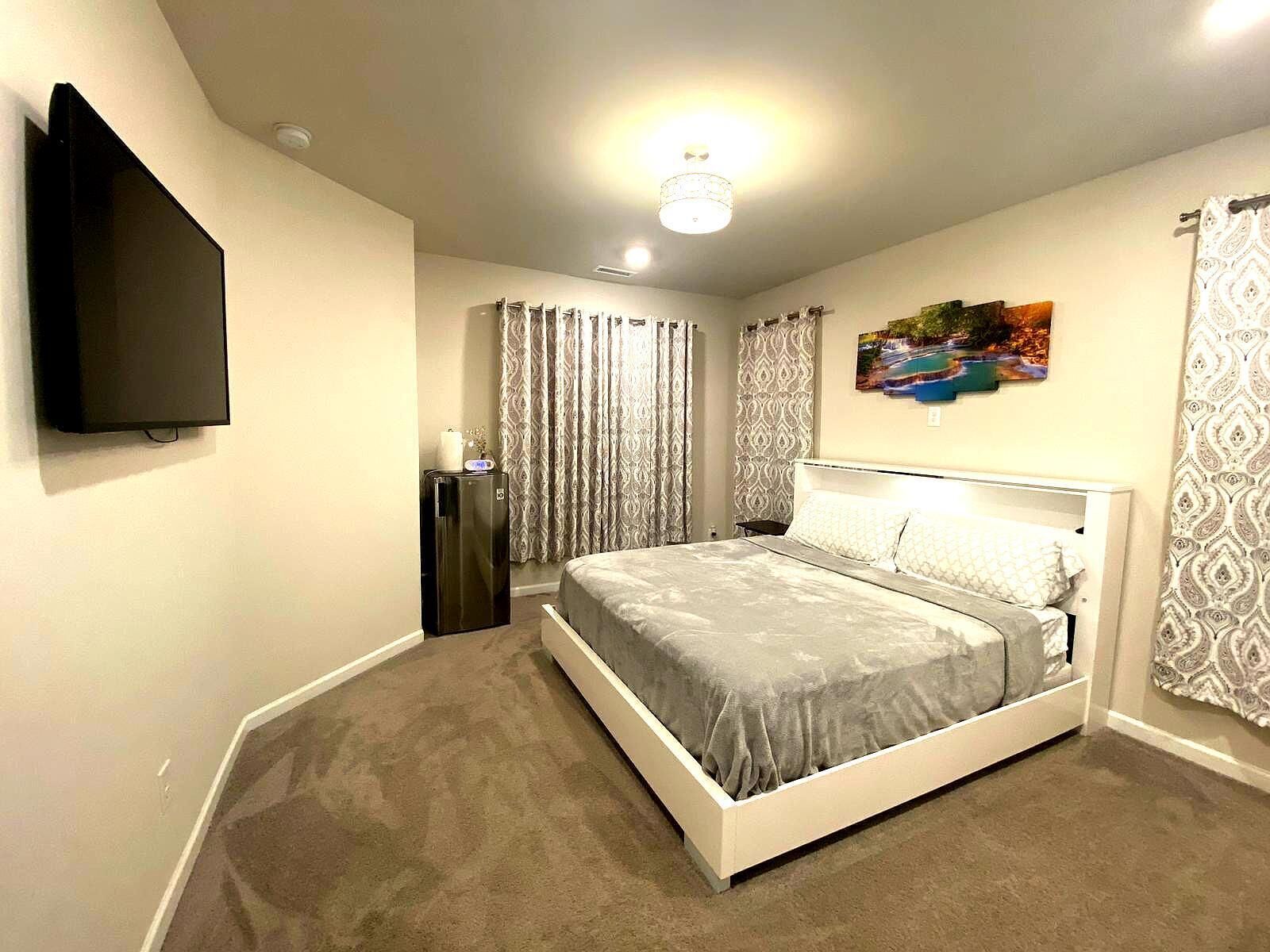 A bedroom with a bed, a flat-screen TV, a refrigerator, and a painting on the wall.