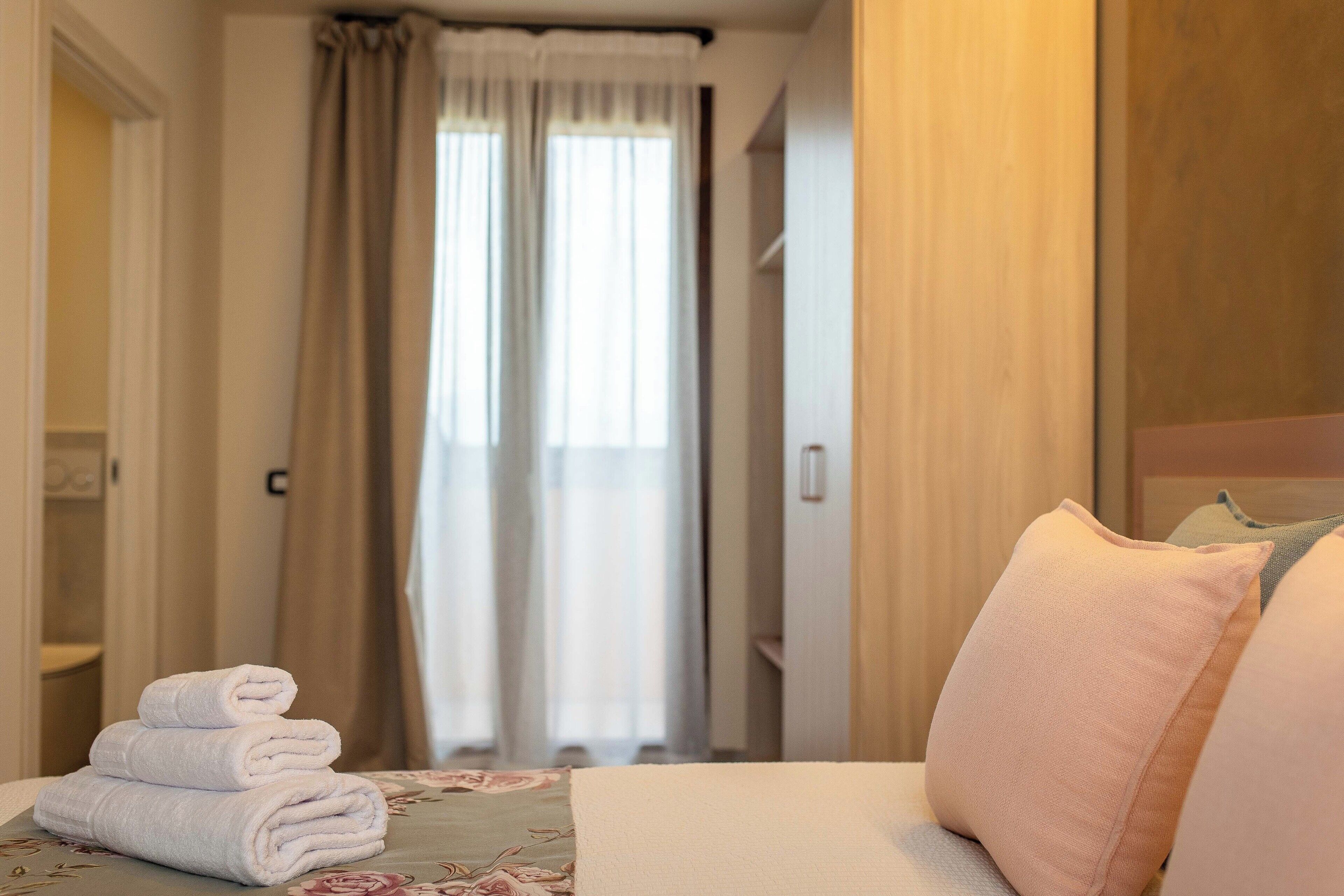 A hotel room with a bed, a stack of folded towels, and a view of the outside through a window with curtains.