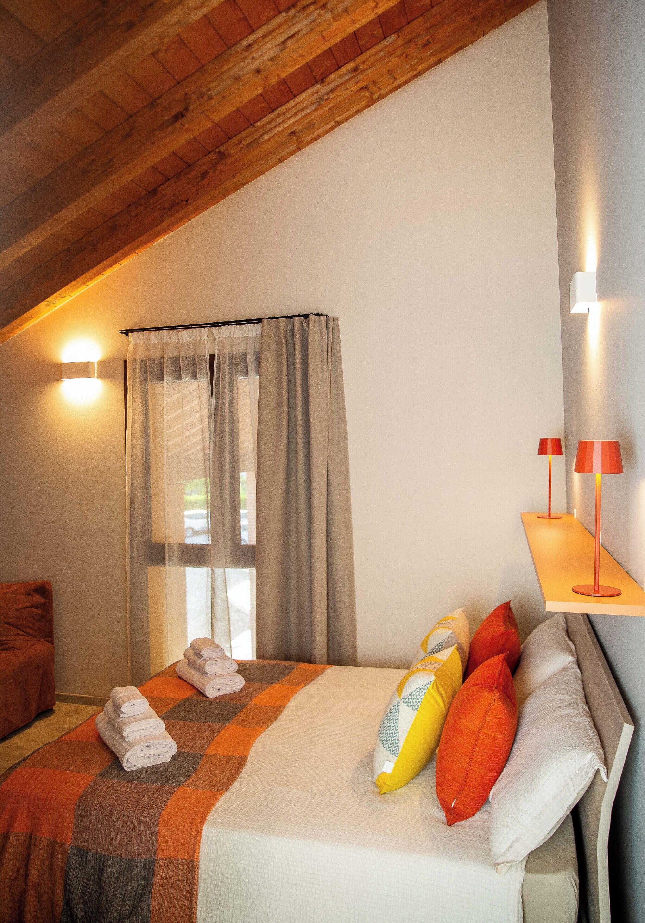 A bedroom with a bed, pillows, a blanket, a window with curtains, a bedside table with lamps, and a wooden ceiling.