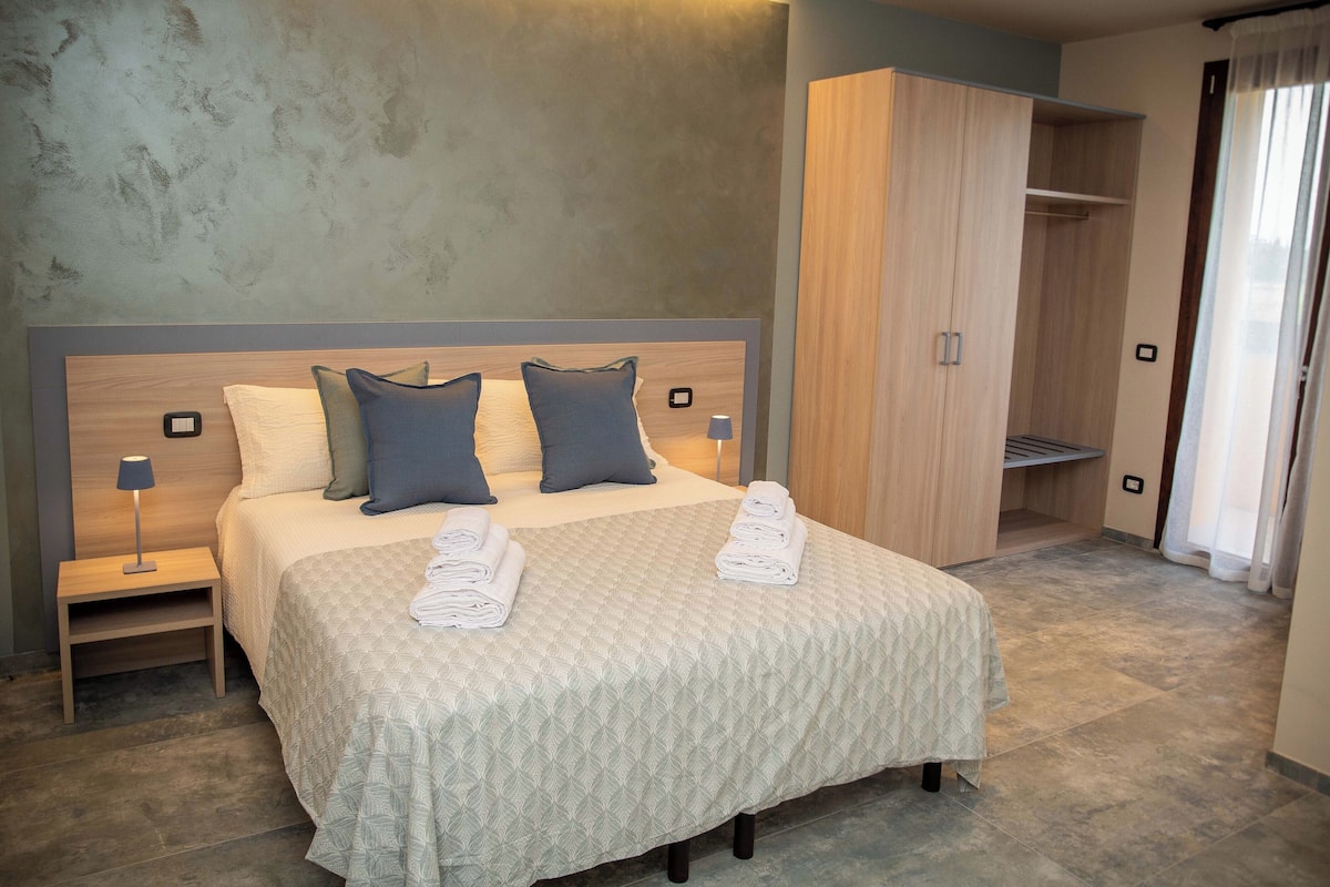 Una cama bien hecha con una colcha acolchada, dos almohadas y toallas dobladas. A la derecha, un armario de madera y, a la izquierda, una mesita de noche con una lámpara.