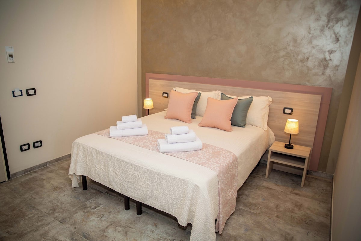 Una cama bien hecha con cabecera, dos mesitas de noche con lámparas y un banco al pie de la cama.