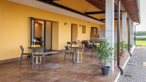 Un'area patio esterna con pavimento piastrellato, una zona salotto coperta con sedie e tavoli, e piante in vaso.