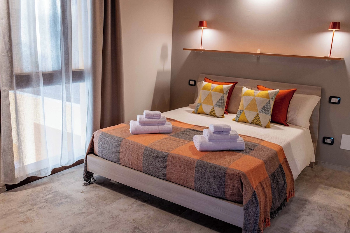 Una cama bien hecha con una colcha de cuadros, dos almohadas con diseños geométricos y toallas dobladas sobre la cama.