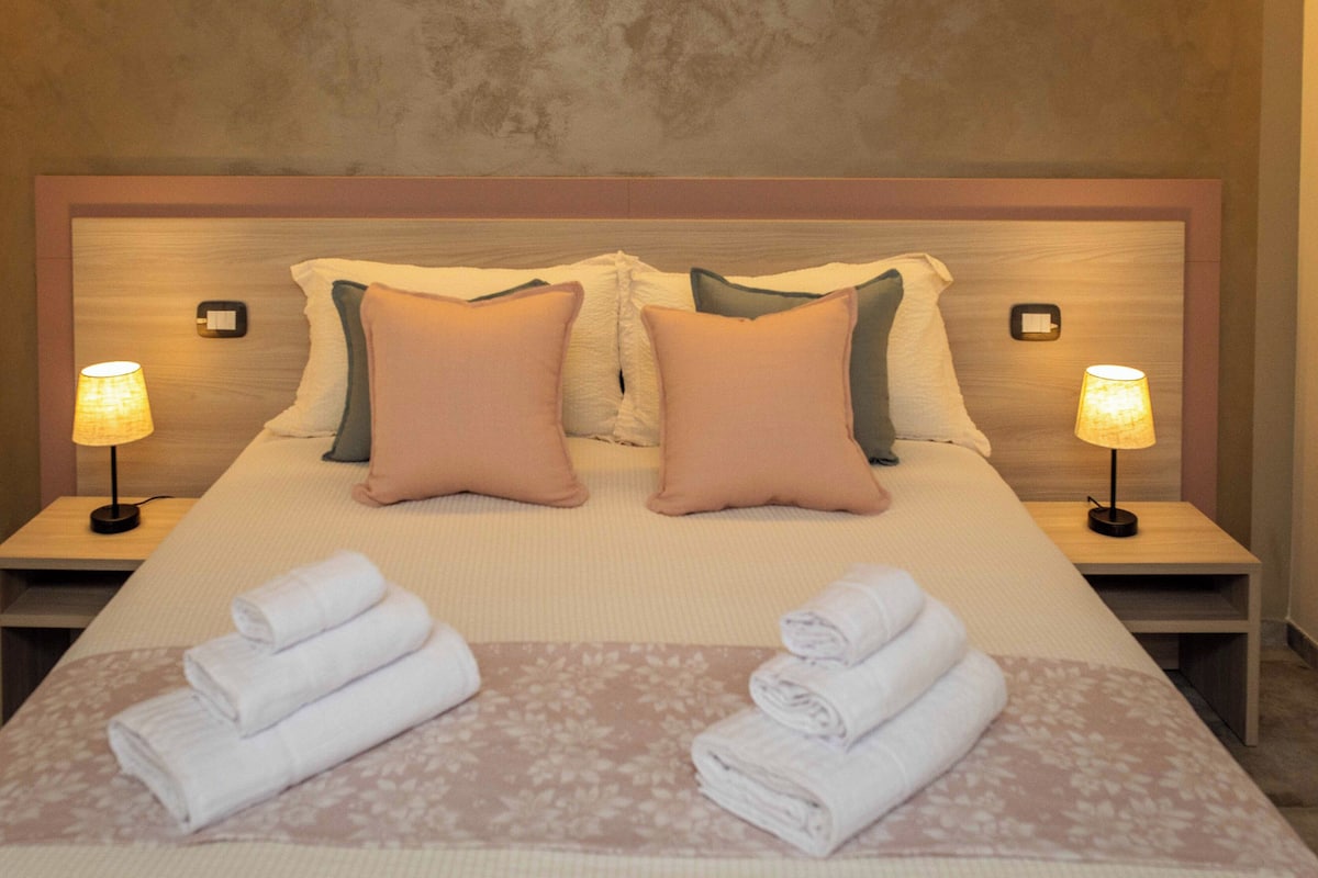 Una cama bien hecha con dos almohadas, dos lámparas de noche y dos toallas dobladas sobre una colcha estampada con motivos florales.