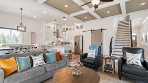 A spacious living area with a gray sofa, black armchairs, and a coffee table. A kitchen is visible in the background, and a staircase leads to another level. - Barefoot Breeze w/Pool, Kayaks, Fireplace (Winchester)