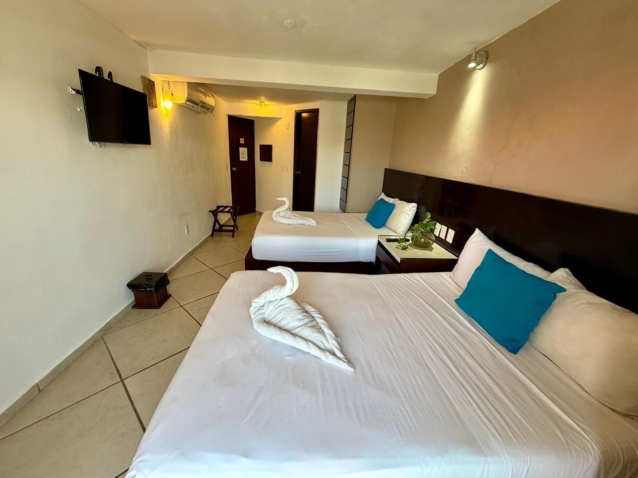 A hotel room with two beds, each with white linens and blue pillows. There is a flat-screen TV mounted on the wall and a small trash bin on the floor.