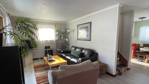 Una sala de estar con un sofá de cuero negro, una mesa de centro de madera y un sillón color crema. Se aprecia una estantería con libros y un globo terráqueo, además de una ventana con cortinas traslúcidas.