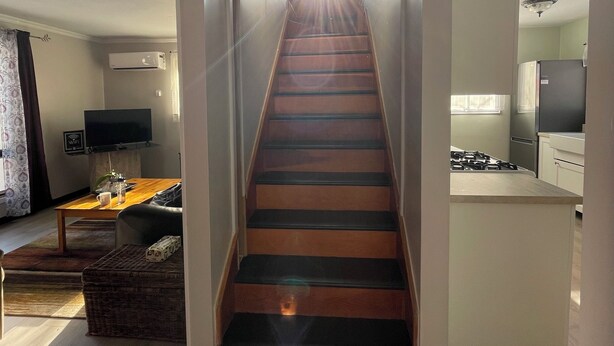 Una escalera conduce a un área de cocina con diseño moderno, un televisor de pantalla plana y un comedor con mesa y sillas.