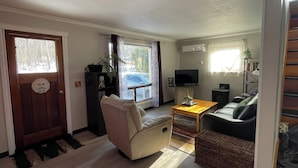 Una sala de estar con un sofá, una mesa de centro y un televisor. Hay una puerta con un tirador decorativo y una ventana con cortinas.