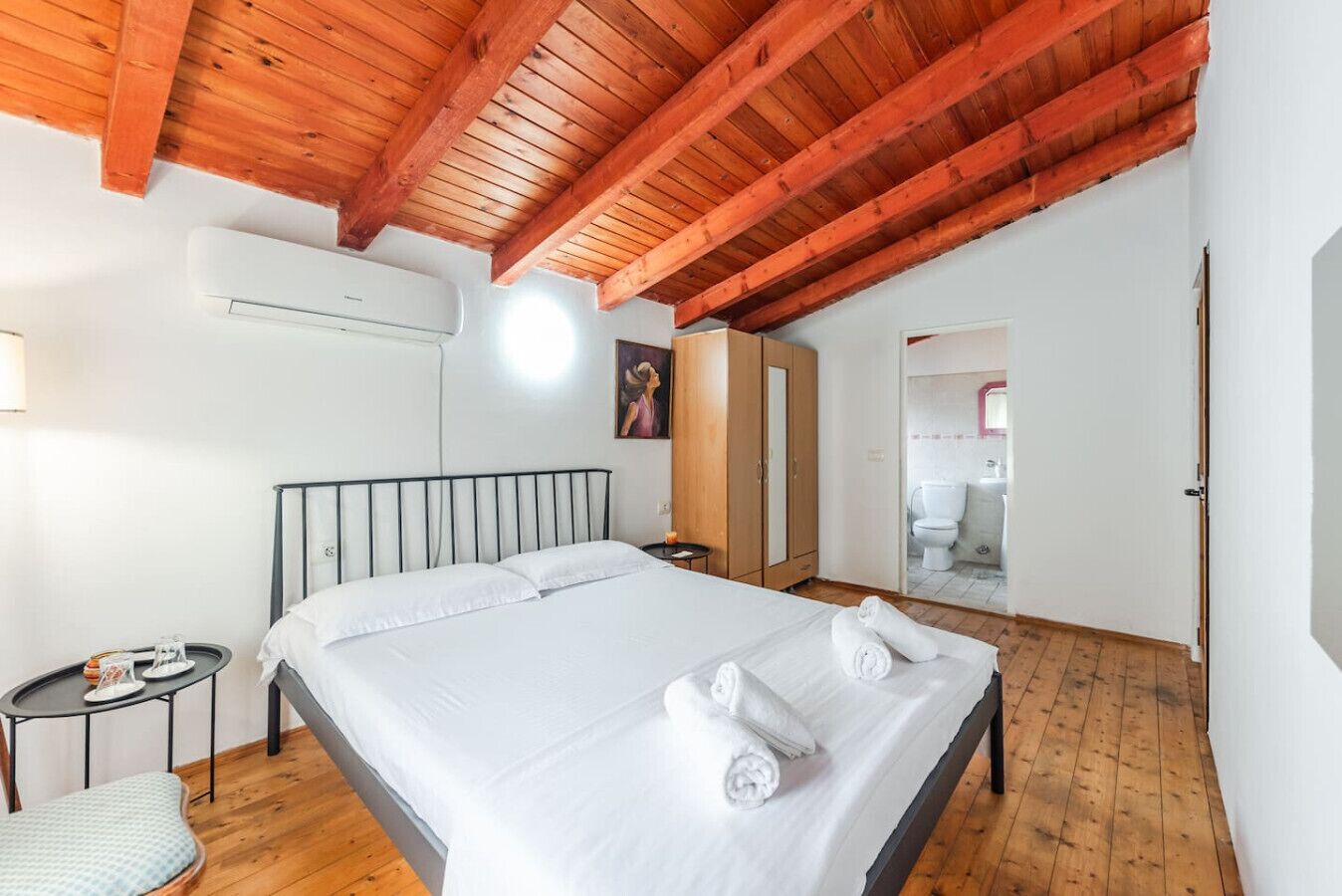 A bedroom with a wooden ceiling, a bed with white linens, a bedside table with a lamp, and a view of the bathroom.