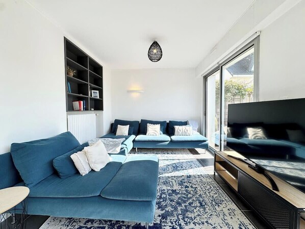 Un salón moderno con un sofá azul, un televisor de pantalla plana y un amplio ventanal que proporciona luz natural.