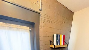Una habitación con una puerta oscura, una cortina blanca, un muro de ladrillo, un estante con toallas a rayas y una rejilla de ventilación.