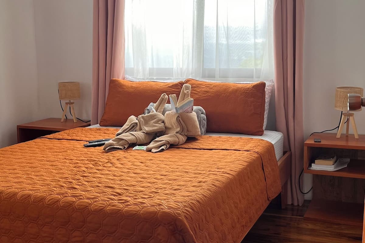 Ein Schlafzimmer mit einem großen Bett, Holzboden, einem Nachttisch mit Lampe und einem Fenster mit Vorhängen.