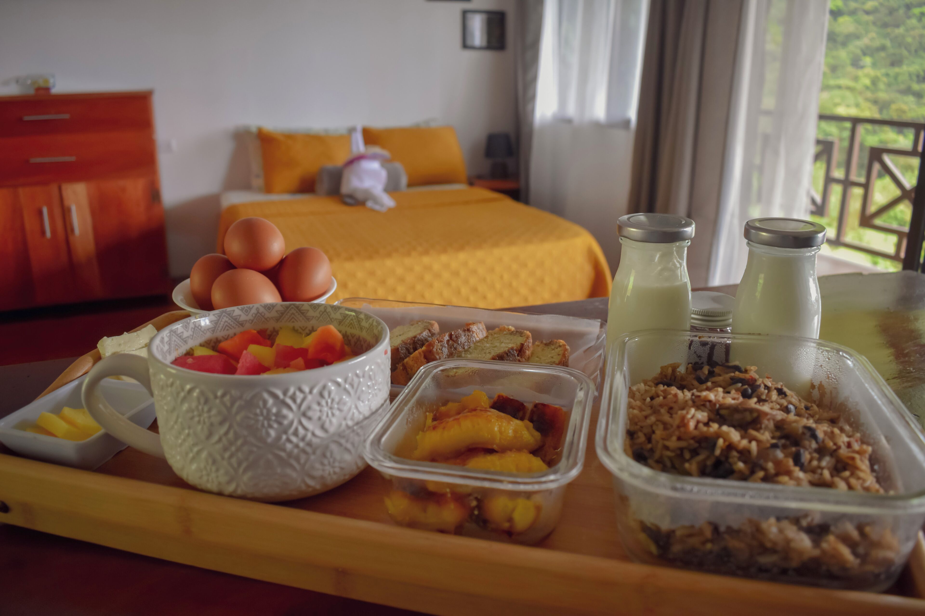 A breakfast tray with eggs, fruit, and cereal in a hotel room with a bed and a view of greenery.
