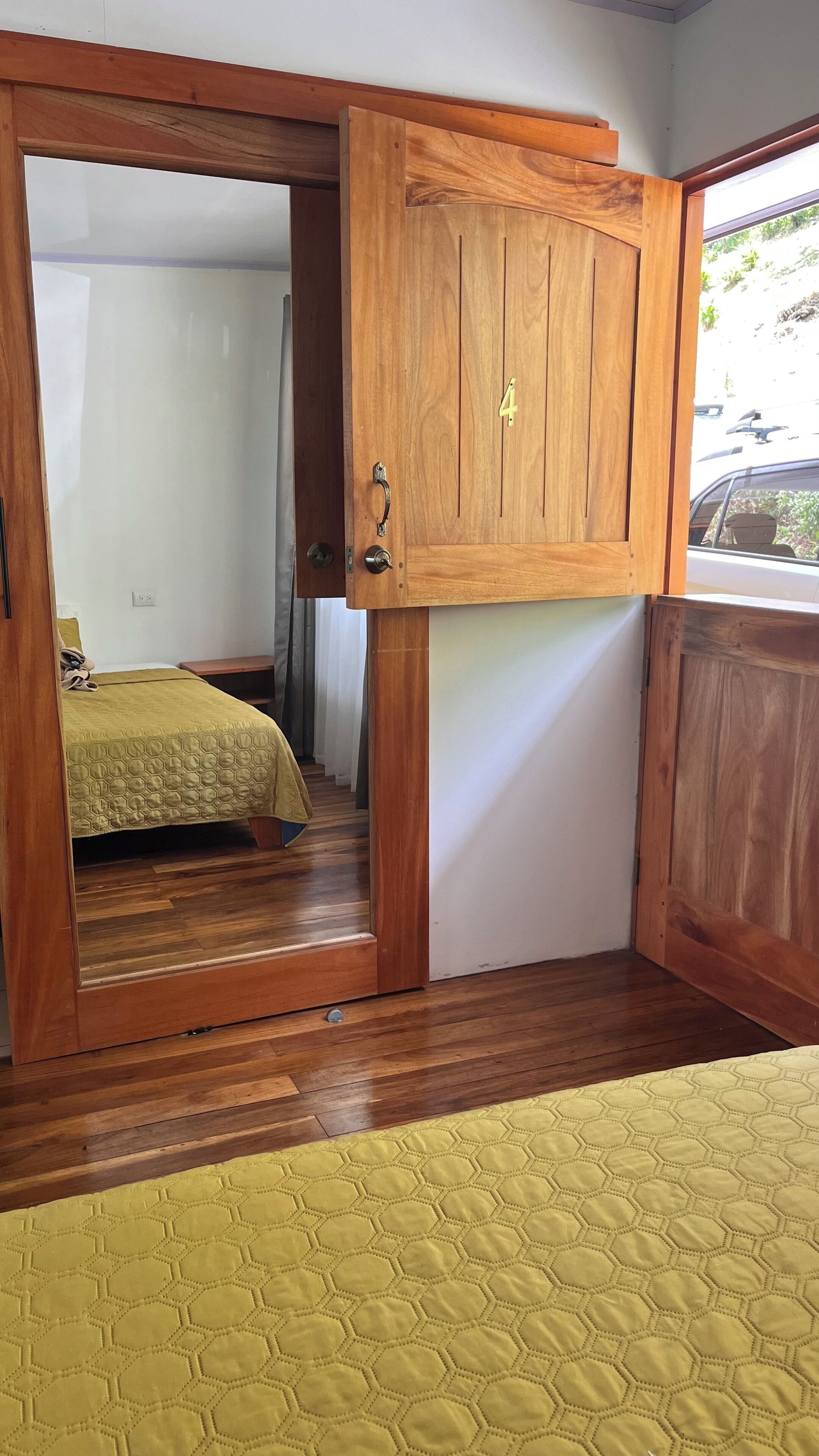 Una habitación con una puerta de madera marcada con el número 4, una cama con colcha amarilla y vista a un coche estacionado afuera.