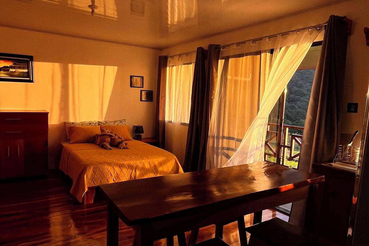 Ein Zimmer mit einem Bett, einem Tisch und einem Stuhl. Es gibt ein Fenster mit Vorhängen.