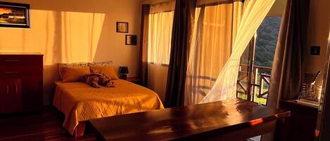 A room with a bed, a table, and a chair. There is a window with curtains.