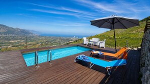 A pool with a view of the sea and mountains, wooden deck, lounge chairs, and a table.