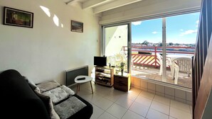A living room with a sofa, a small table, a TV, and a sliding glass door leading to a balcony with a view of a parking area and buildings. - Bleu Courant - T4 current view (Mimizan)