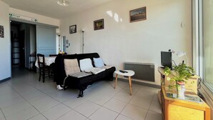 A living room with a black sofa, a small round table, a radiator, and a TV on a wooden stand. - Bleu Courant - T4 current view (Mimizan)
