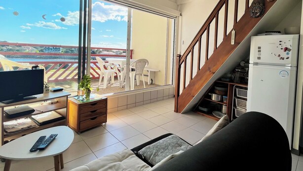 A living room with a TV, a sofa, a small table, and a balcony with a view of the sea and buildings. - Bleu Courant - T4 current view (Mimizan)