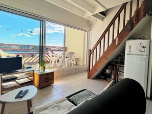 A living room with a TV, a sofa, a small table, and a balcony with a view of the sea and buildings. - Bleu Courant - T4 current view (Mimizan)
