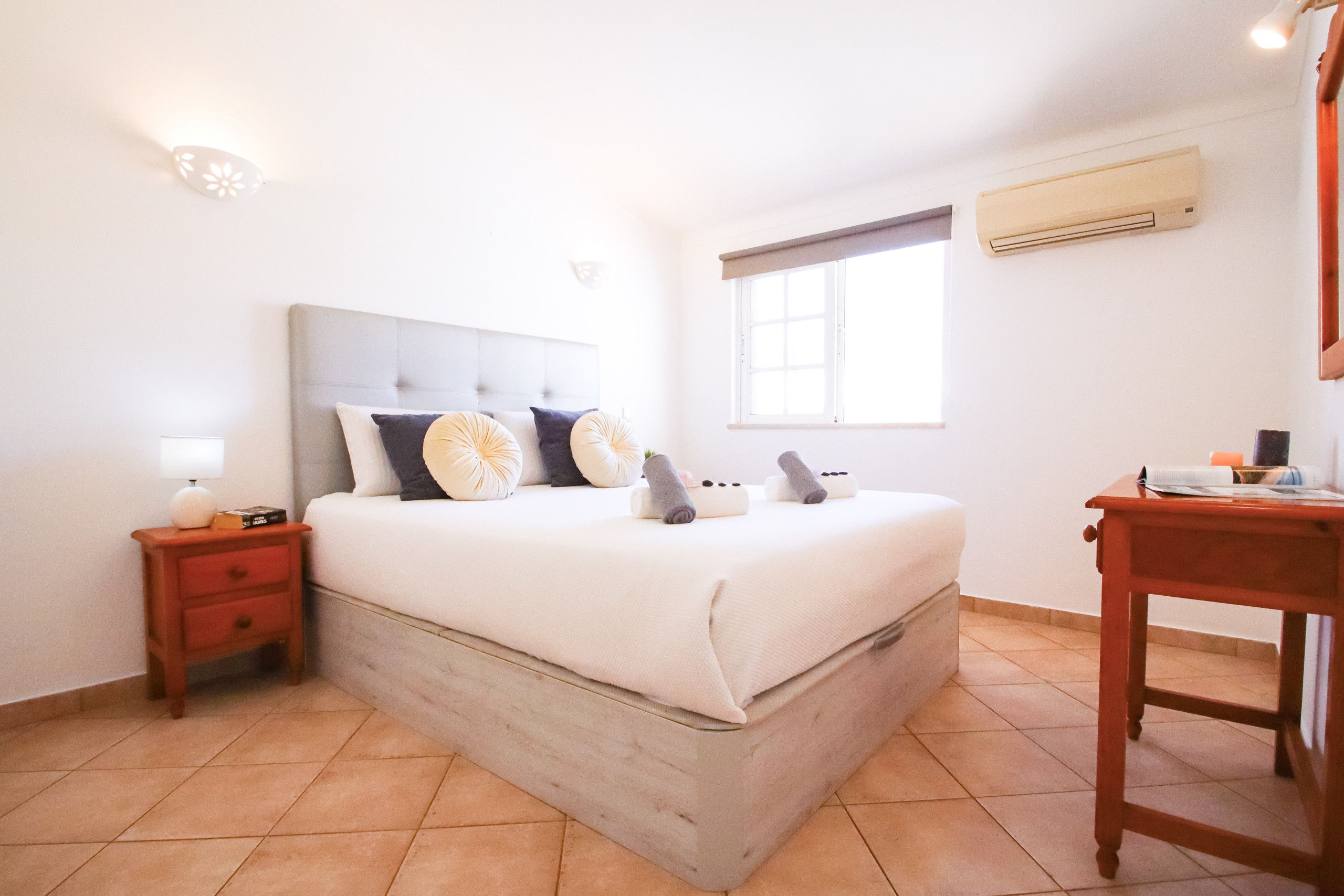 A bedroom with a bed, nightstand, lamp, and a window with an air conditioning unit.