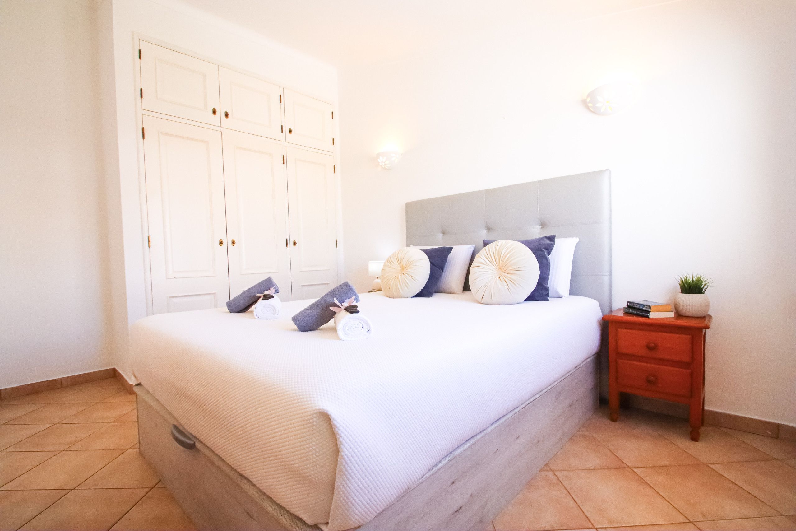 A bedroom with a bed, pillows, a nightstand, a potted plant, and a wardrobe.