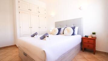 A bedroom with a bed, pillows, a nightstand, a potted plant, and a wardrobe.