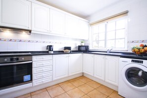Una cocina con armarios blancos, encimera negra, microondas, lavadora y una ventana con persianas.