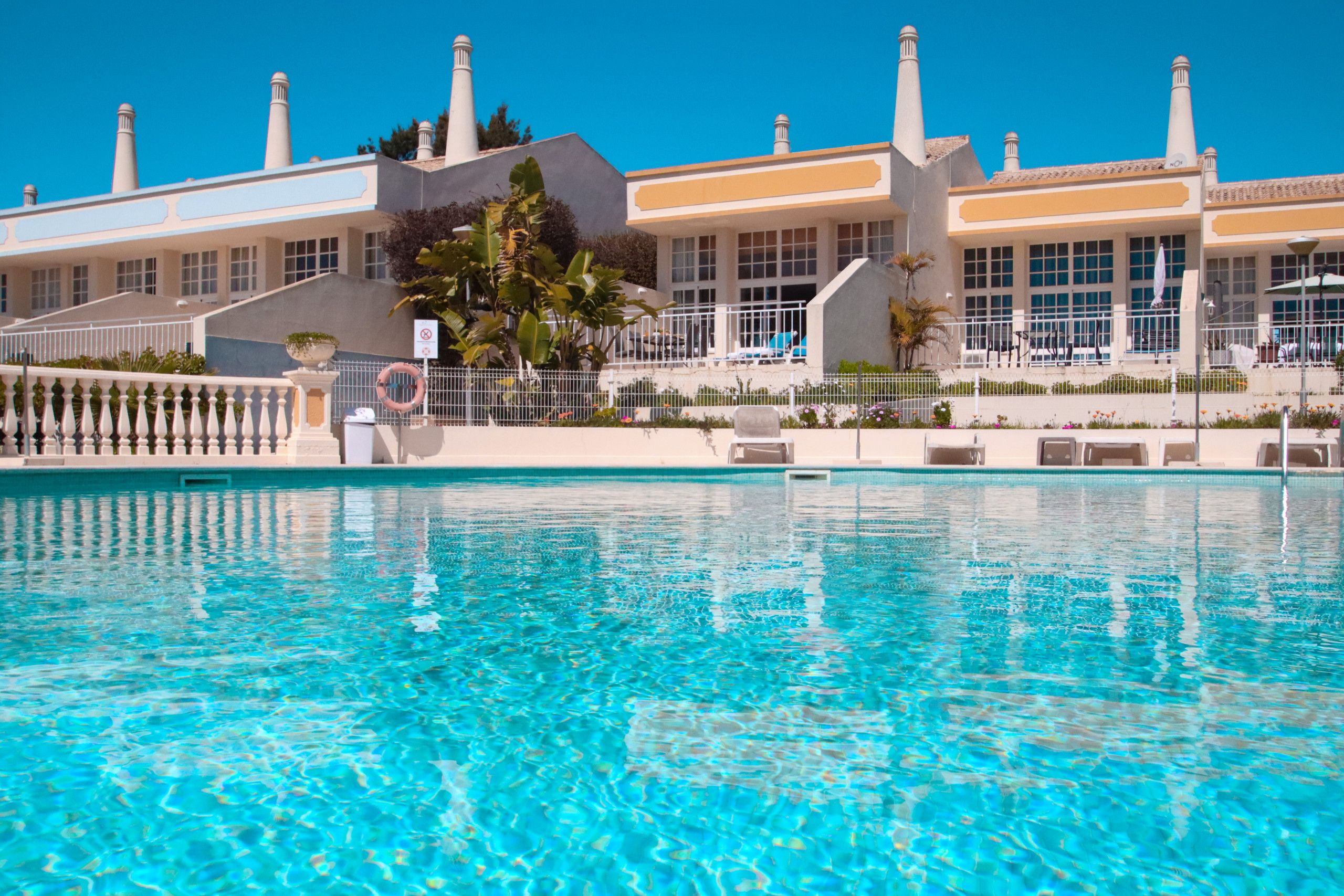 Une piscine à l’eau d’un bleu cristallin, entourée d’une clôture blanche et bordée d’un bâtiment doté de nombreuses cheminées et de grandes fenêtres.