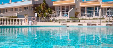 Uma piscina com superfície de água azul cristalina, cercada por uma cerca branca e um edifício com várias chaminés e grandes janelas.