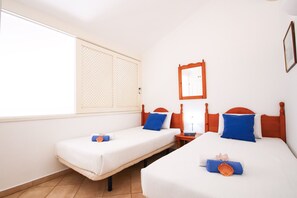 Une chambre d’hôtel avec deux lits simples, une fenêtre avec un store blanc, une table de chevet avec une lampe et un miroir fixé au mur.