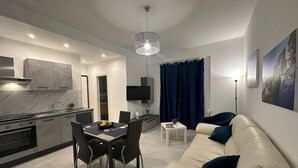 A modern living room with a beige sofa, a dining table with chairs, a kitchen area with a stove and cabinets, a TV mounted on the wall, and a large window with curtains. - Blue Heracleo Apartment center Trapani (Trapani)