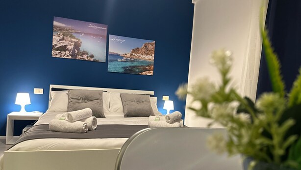 A hotel room with a bed, two bedside lamps, a nightstand, and two framed pictures on the wall. - Blue Heracleo Apartment center Trapani (Trapani)