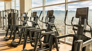 Un gimnasio con varias máquinas de ejercicio, ventanas amplias y vistas a un paisaje urbano.