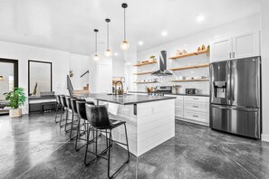 A modern kitchen with a central island, bar stools, stainless steel appliances, and open shelving.