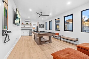 A modern living room with a pool table, a ceiling fan, and large windows offering views of the outside.