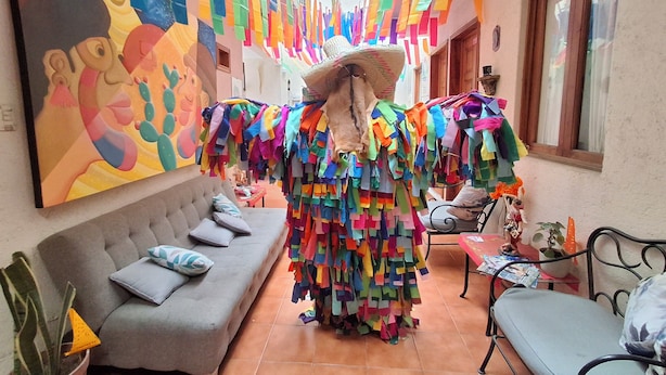 Une pièce décorée de motifs colorés en papier suspendus au plafond, un canapé gris avec des coussins, une petite table avec une plante et une fenêtre avec des rideaux.