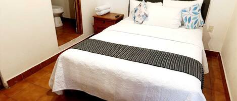 A neatly made bed with a white bedspread and a black and white striped runner, a wooden nightstand with a folded white towel, and a bathroom visible through an open door.