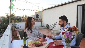 Un couple est assis à une table en plein air, recouverte d’une nappe blanche, avec une bouteille de vin et une variété d’amuse-gueules.